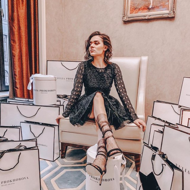 Аделина Сотникова в черном платье сидит на белом кресле в окружении подарков