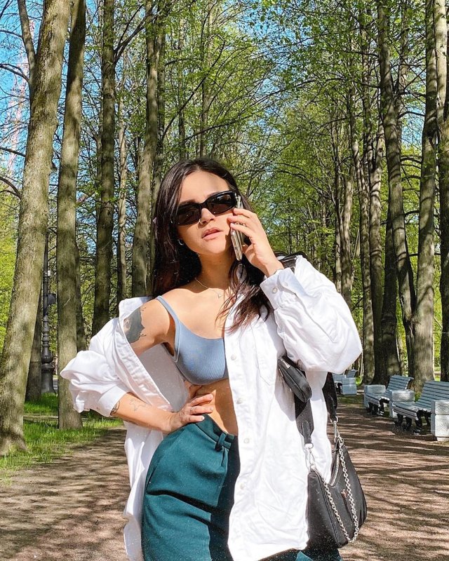 Ирина Смелая (Tatarka) в парке в джинсах, белой рубашке и сером топе разговаривает по телефону