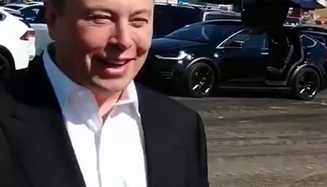 Илон Маск в черном пиджаке и белой рубашке на фоне Tesla