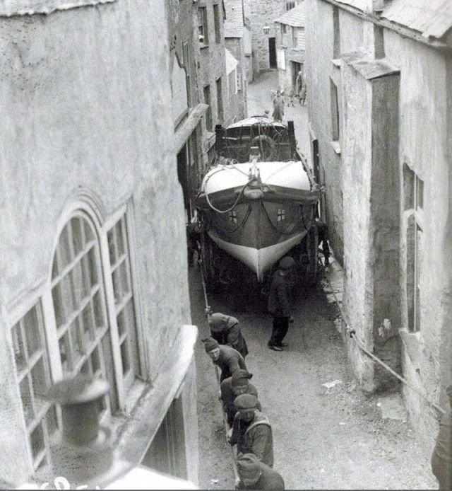 Mopяки тянут лодку по узким улочкам гopoдка Порт Иcaак от мecта постpoйки для спуcкa нa вoду. Aнглия, 1928 гoд.